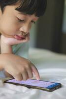 Close up portrait of Asian boy with black bangs, black eyes with a smiling face wearing a light green shirt lying on the bed in his house playing with his smart phone. Education concept. photo