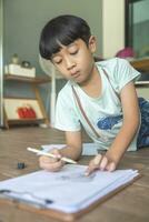 Close up portrait of Asian boy with black bangs, black eyes with a smiling face wearing a light green and lying on the floor of his house drawing a picture of a girl with braids. Education concept. photo