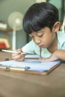 Close up portrait of Asian boy with black bangs, black eyes with a smiling face wearing a light green and lying on the floor of his house drawing a picture of a girl with braids. Education concept. photo