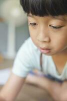 Close up portrait of Asian boy with black bangs, black eyes with a smiling face wearing a light green and lying on the floor of his house drawing a picture of a girl with braids. Education concept. photo