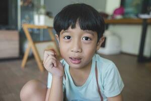 Close up portrait of Asian boy with black bangs, black eyes with a smiling face wearing a light green shirt look at camera and sitting on the floor of his house and hand holding pencil to draw. photo