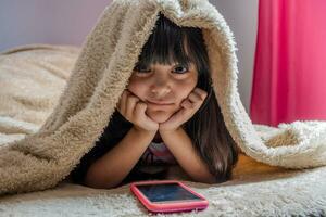 un niño utilizando un teléfono inteligente acostado en cama, jugando juegos, acecho en línea vídeos, desplazamiento el pantalla. para niños pantalla adiccion. para niños habitación. foto