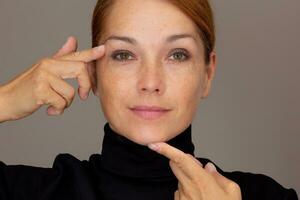 Portrait of cropped caucasian smiling middle aged woman face with freckles showing on eyelid and chin by index fingers photo