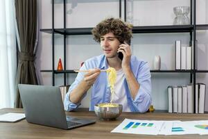 caucásico hombre es trabajando a su hogar oficina respondiendo el teléfono llamada desde cliente mientras comiendo instante fideos y analizando el estadística gráfico para Lanza libre y empresario negocio concepto foto