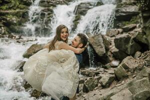 Portrait of a happy bride and groom near a waterfall. The groom holds the bride in his arms. Wedding photo session in nature. Photo session in the forest of the bride and groom.
