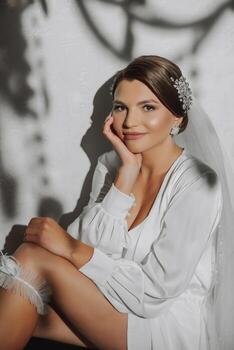 A young bride in a white robe with a wedding garter on her leg, close-up. Preparation for the wedding ceremony. Accessories for newlyweds, lace garter. photo
