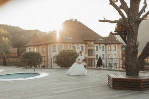 A groom in a black suit and a blonde bride dance a waltz against the background of a mountain and buildings, near a swimming pool. Long dress in the air. Romantic photo in nature. Beautiful hair