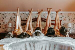 Beautiful sexy girls best friends wearing robes lying on bed in a row raise legs up celebrate bridal shower bachelorette spa party together epilation waxing legs skin beauty concept photo