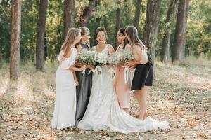 Boda foto en naturaleza. el novia y su damas de honor son en pie en el bosque sonriente, participación su ramo de flores y mirando a el novia. contento Boda concepto. emociones muchachas