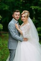 Portrait of the bride and groom standing against the background of green trees, hugging and looking into the lens. Stylish groom. Fashion and style. Beautiful bride photo