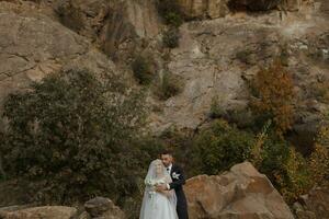 The bride and groom are standing on a rock high in the mountains next to a large stone. The bride is under a veil, the groom is hugging her shoulders photo