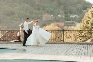 A groom in a black suit and a blonde bride dance a waltz against the background of a mountain and buildings, near a swimming pool. Long dress in the air. Romantic photo in nature. Beautiful hair