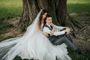 un hermosa novia con largo Rizado pelo en un elegante vestir abrazos el acicalar, sonrisas, mirando dentro el lente debajo un grande árbol. retrato de el novia y novio. primavera boda. natural maquillaje foto