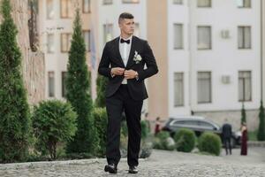 A man in a white shirt, a black bow tie and a black suit holds a bouquet and goes to a meeting, behind him are black cars, new buildings and trees. A stylish watch. Men's style. Fashion. Business photo