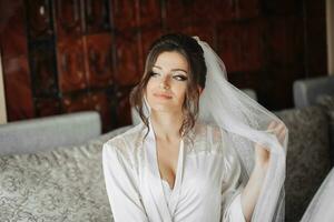 Portrait of the bride. A brunette bride is sitting on a gray sofa in a dressing gown, posing, holding her voluminous white veil. Gorgeous make-up and hair. Wedding photo. Beautiful bride photo
