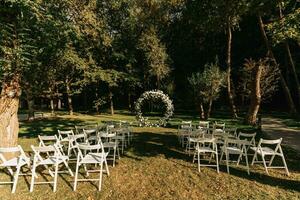 Wedding ceremony in the forest. The road to the round arch with foliage, greens, greenery, and flowers. Rustic decor. Wooden chairs in the backyard banquet area. Seats for guests. photo