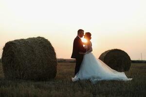Wedding portrait of the bride and groom. Bride and groom stand embracing at sunset near hay bales. Red-haired bride in a long dress. Stylish, bearded groom. Silhouette photo. photo