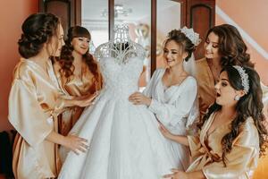 Young bridesmaids in silk robes in a hotel room. Beautiful women celebrate a bachelorette party, next to a luxurious dress of the bride in themed pajamas. photo