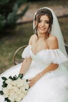 Portrait of the bride in nature. Brunette bride in a white long dress, holding a bouquet of white roses, sitting on a bench, posing, looking down, against a tree background. Curly hair photo