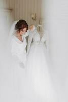 un hermosa joven novia en un blanco enaguas en pie cerca su Boda vestido. final preparativos para el boda. el novia es esperando para el novio. mañana, novia. foto