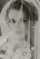 Curly blonde bride in a white dress, covered with a veil, poses for the camera with a bouquet of roses. Portrait of the bride. Beautiful makeup and hair. Wedding in nature. black and white photo