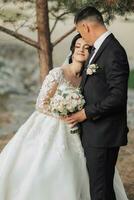 Wedding portrait in nature. The brunette bride and groom in a white long dress are standing and hugging against the background of coniferous trees and a white hut. photo