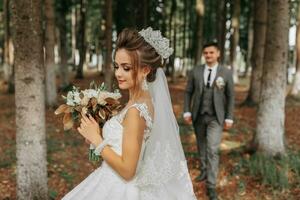 A beautiful young woman in a wedding dress between tall trees in the forest with a royal hairstyle and a chic tiara with a bouquet of flowers in her hands, a wedding in golden color photo