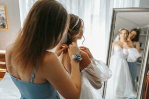 The bride's friend helps in the morning, fastens a chain with a pendant with a boutonniere on her hand, the bride has a beautiful hairstyle and light natural makeup. Wedding portrait. photo