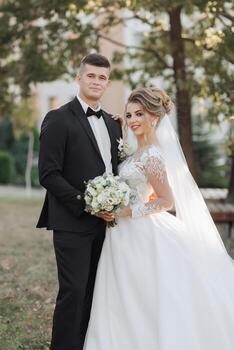 Wedding portrait. The groom in a black suit and the blonde bride are standing, hugging, posing holding a bouquet under a tree. Photo session in nature. Beautiful hair and makeup