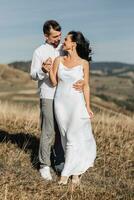 un elegante modelo Pareja en el montañas en el verano. un joven chico y un niña en un blanco seda vestir son caminando en el Pendiente en contra el antecedentes de el bosque y montaña picos chico Besos un niña foto