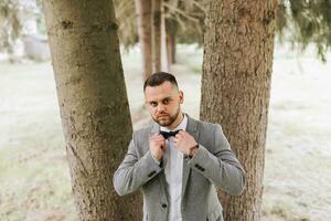 stylish groom, leaning his shoulder against a tree, emphasizes his beard, looks into the camera lens. Portrait photo