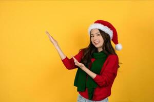 Asian woman wearing red sweater, scarf and santa hat with arms raised to empty space aside on a yellow studio background. photo