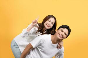 Happy Asian couple in white casual clothes enjoying piggyback ride and pointing at empty space on yellow background. photo