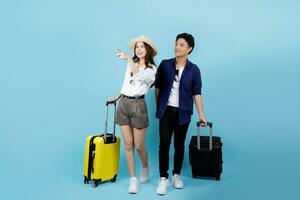 Happy Asian couple tourist hand pointing to copy space with suitcases going to travel on holidays isolated on blue background. photo