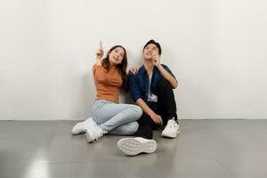 Portrait of cheerful young Asian couple in casual outfits sitting on floor and pointing up offering space for your ad design photo