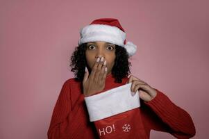 Surprised dark skinned santa girl with stocking on pink background.Merry christmas and winter holidays concept photo