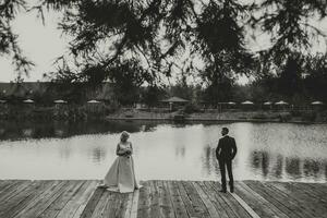 el novio en un negro traje y blanco camisa soportes a un distancia desde el novia en un blanco Boda vestido, en pie cerca un lago y rocas foto