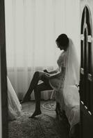 A brunette bride is sitting, posing, dressed in a robe and a long veil, and wearing a wedding band on her leg. wedding portrait, wedding black and white photo. Beautiful makeup and hair. photo