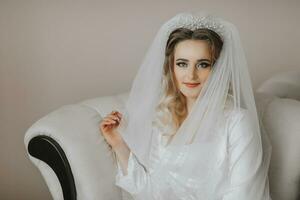 The blonde bride is dressed in a satin robe of seats and a sofa under a veil. Beautiful hair and make-up, open bust. Wedding portrait. Sincere smile. photo