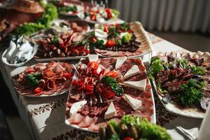 Buffet table at the wedding. An assortment of snacks on white plates standing on a Ukrainian-style tablecloth. Banquet service. Food, snacks with cheese, ham, prosciutto and greens. photo