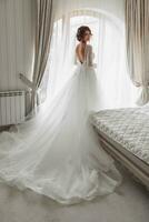 el novia en un hermosa vestir es en pie por el ventana con un largo tren. boda. Boda ramo de flores Mañana de el novia. un contento novia es esperando para el novio. foto