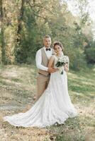 Wedding photo in nature. The bride and groom are standing in the forest, smiling and looking at the camera. The groom hugs his beloved from behind, the bride holds a bouquet. Portrait. Summer wedding