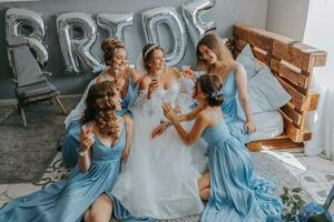 joven damas de honor en azul seda vestidos bebida champán en el de la novia habitación. hermosa mujer celebrando despedida de soltera fiesta sentado en cama y con champán. foto