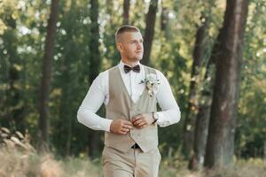 retrato de el novio en el bosque. el novio ajusta su ligero gris chaleco. elegante, elegante novio posando con marrón arco atar, mirar, chaleco y blanco camisa. foto