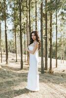 un niña con largo negro Rizado pelo soplo en el viento es en pie en un blanco seda vestido. en pie en el primavera bosque en contra el antecedentes de grande arboles foto