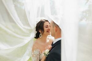 A curly-haired brunette bride in a white dress and a veiled groom embrace and kiss. Portrait of the bride and groom. Beautiful makeup and hair. Wedding in nature photo