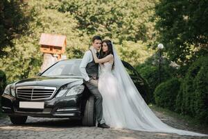 gorgeous bride with trendy makeup and hairdo in luxury wedding dress with handsome groom near black wedding car photo