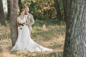 Wedding photo in nature. The bride and groom are standing near a tree smiling and looking at the camera. The groom hugs his beloved from behind, the bride holds a bouquet. Portrait. Summer wedding