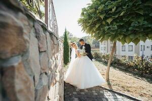 Wedding portrait. The groom in a black suit and the blonde bride stand by a stone wall under a tree, the groom kisses the bride's hand. Photo session in nature. Beautiful hair and makeup