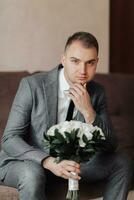 Portrait. A man in a white shirt, black tie and gray suit is sitting on a sofa, holding a bouquet of white roses. A stylish watch. Men's style. Fashion. Business photo